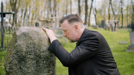 Close-Up-View-Of-Man-In-Black-Suit-Kneeling-In-Front-Of-A-Grave