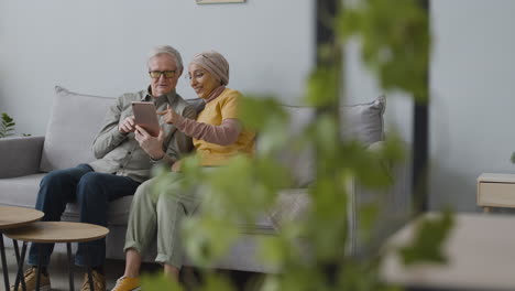 Mujer-árabe-Enseñando-A-Un-Anciano-A-Usar-Una-Tableta-Mientras-Están-Sentados-En-Un-Sofá-En-Casa