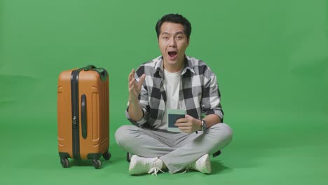 짐과 여권과 함께 아시아 남성 여행자의 몸이 가득 차 있으며, 녹색 스크린 배경 스튜디오에 앉아있는 동안 웃고 와우라고 말합니다.