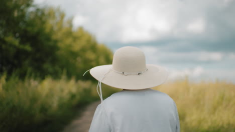 back view of woman wearing wide-brimmed hat and cyan shirt walking along dirt path surrounded by lush greenery under partly cloudy sky, enjoying peaceful countryside stroll