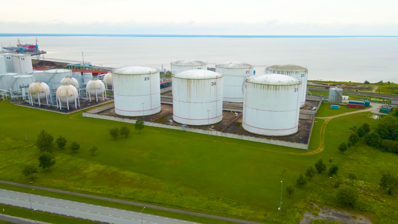 Aerial drone footage of large white tanks for liquid storage near a cloudy and misty port or harbor where containers, railroad and highway is visible. Storaging gas and oil for global logistics trade