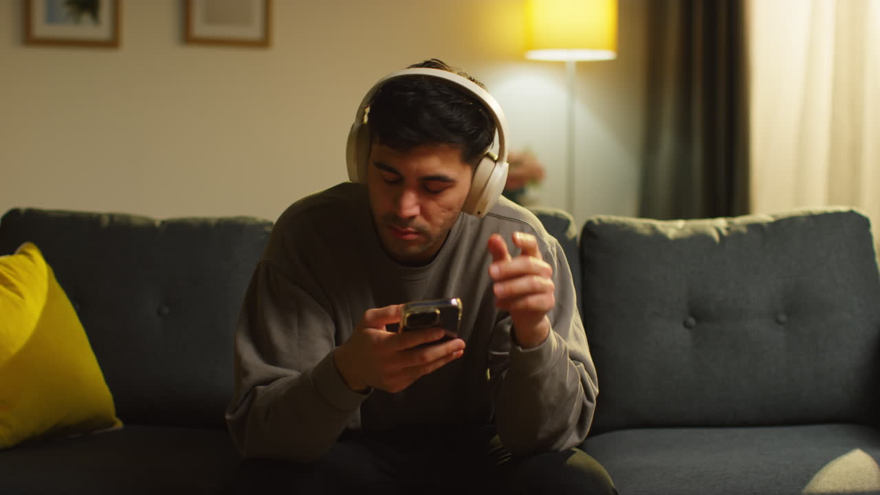 무선 헤드폰을 착용하여 소파에 앉아 휴대 전화로 음악을 스트리밍하는 젊은이가 집에서 저녁을 보내고 있습니다.