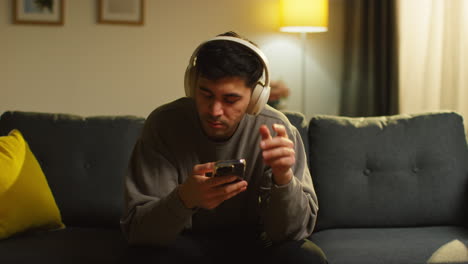 joven pasando la noche en casa sentado en el sofá con auriculares inalámbricos transmitiendo música desde el teléfono móvil contestando llamadas