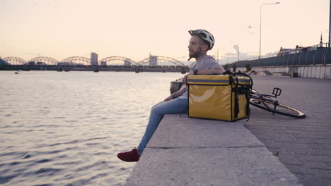 food delivery man rests at sunset next to his thermal backpack looking at the beautiful scenery on the shore of a river