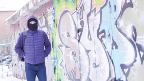 man in hooded jacket and mask walking on a snowy street by a graffiti wall