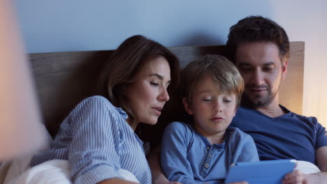 vista de cerca de los padres caucásicos sentados en la cama con su pequeño hijo lindo, viendo algo en la tableta y hablando por la noche