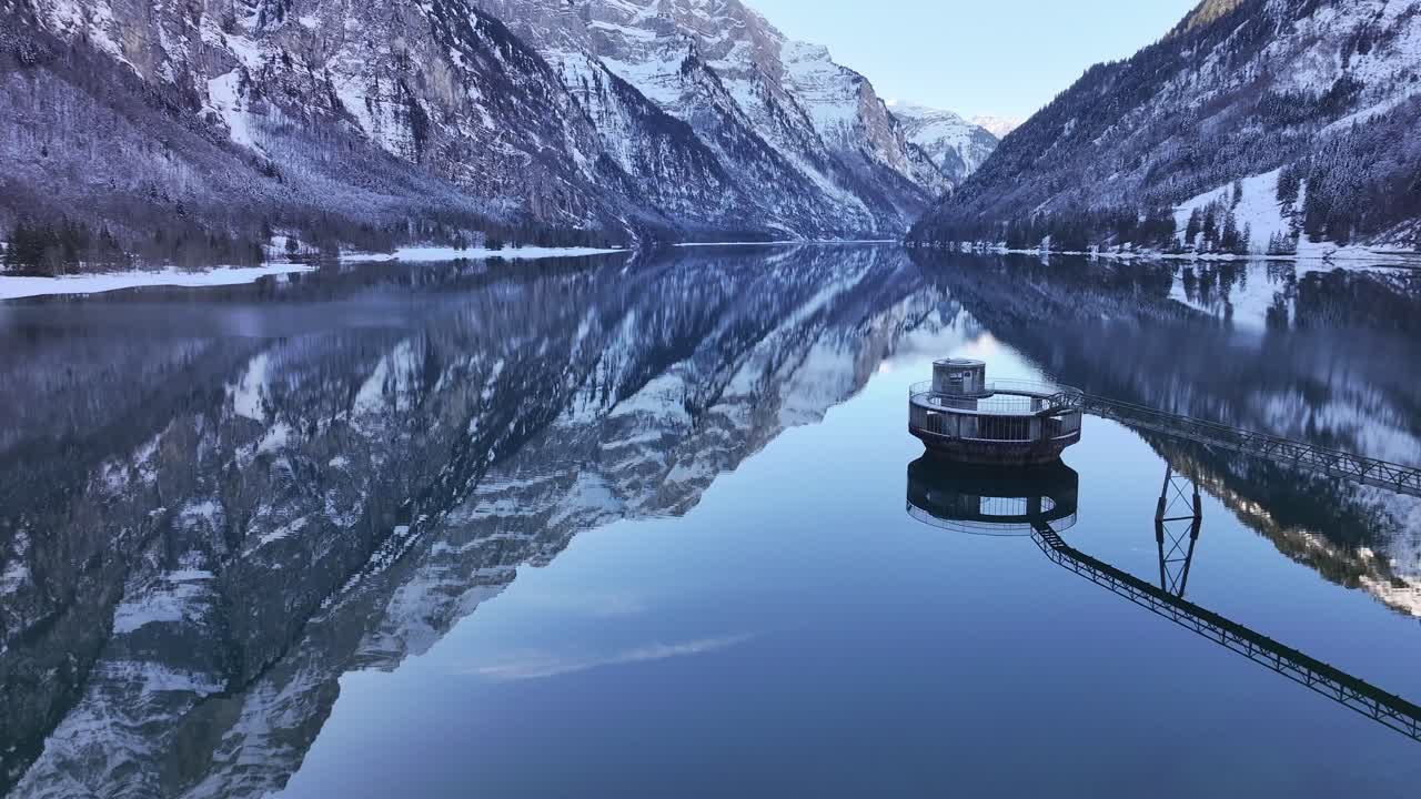 A captivating view of the Klöntalersee in Switzerland during winter. The serene lake, nestled amidst snow-capped mountains, reflects their majestic peaks in a mirror-like image.