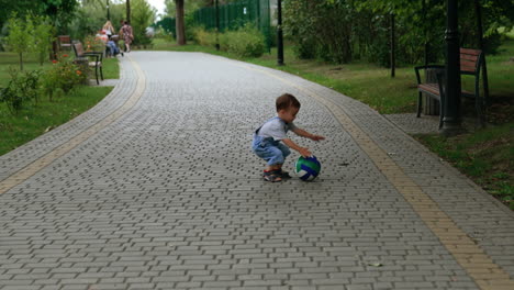 kid runs by the paved road to catch up his ball. cute toddler squats to pick up his toy.