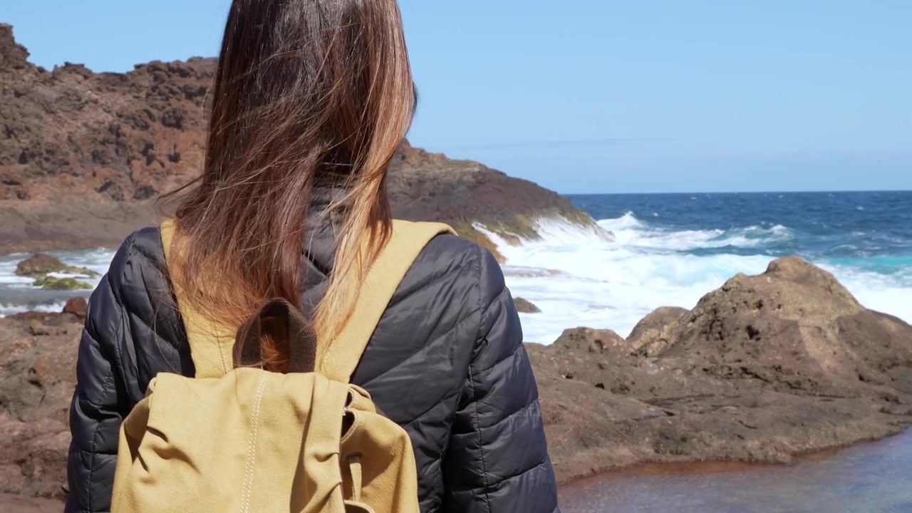 Woman with a backpack standing on a rocky volcanic coast. Brave female explorer enjoying the beautiful scenery during her trip