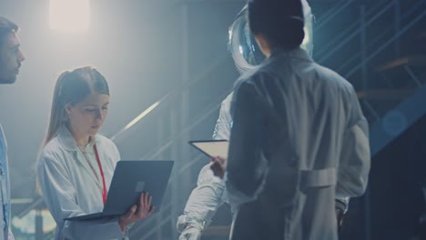 백색 코트를 입은 다양한 항공우주 과학자 및 엔지니어 팀이 토론하고 컴퓨터를 사용하며 은하 탐사 및 여행에 적응한 새로운 우주복을 설계합니다.
