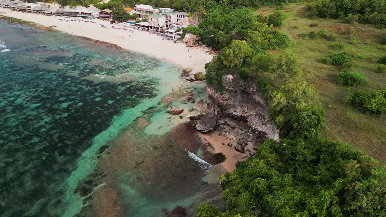 Coastal drone video of Balangan Beach Bali capturing turquoise reef shallows, volcanic cliff formations, sandy bay and resort area along a clear tropical shoreline with blue lagoon water