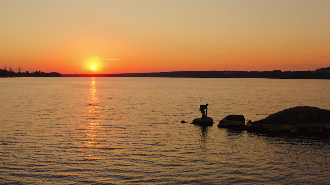 River at sunset. Silhouette of a fisherman on stone. Man going away from the lake after fishing on orange sky background in the evening.