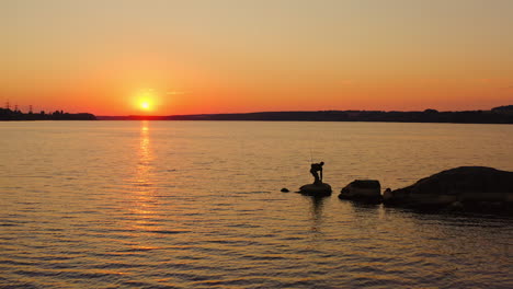 river at sunset. silhouette of a fisherman on stone. man going away from the lake after fishing on orange sky background in the evening.