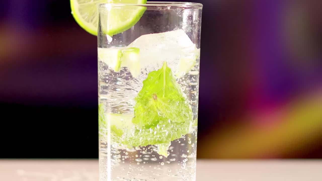 A close-up of a glass with lime, mint, and ice against a vibrant, blurred background.
