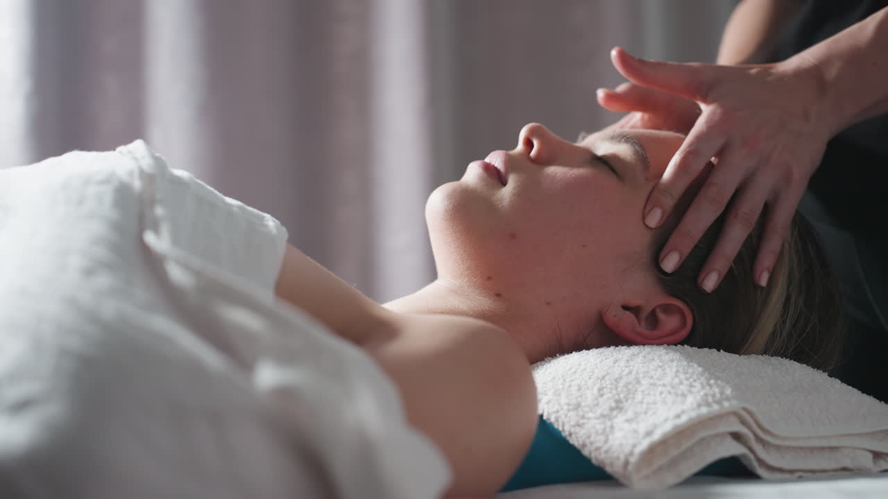 Close up of woman lying peacefully with eyes closed while therapist's hands massage forehead and head, soft light illuminating her face and towel-covered support, creating comforting ambiance