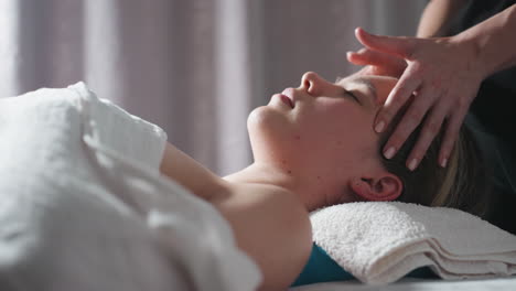 close up of woman lying peacefully with eyes closed while therapist's hands massage forehead and head, soft light illuminating her face and towel-covered support, creating comforting ambiance