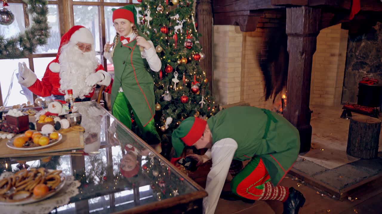 Santa with funny elves at Christmas. Elf is taking photo of Santa and girl elf on Christmas room background. New Year time.