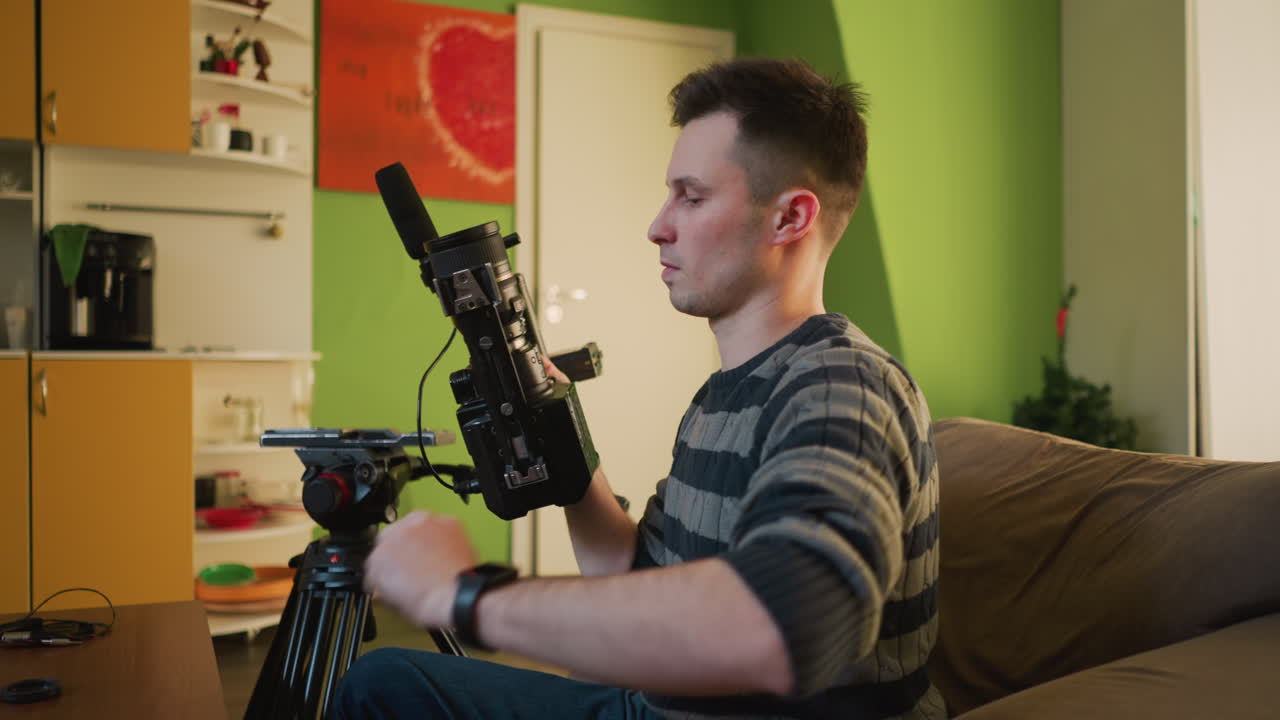 Rapaz do estúdio ajustando a câmera no tripé em estúdio caseiro, preparando-se para uma gravação de vídeo. Câmera, microfone e equipamento de iluminação montados na mesa, preparando-se para a produção