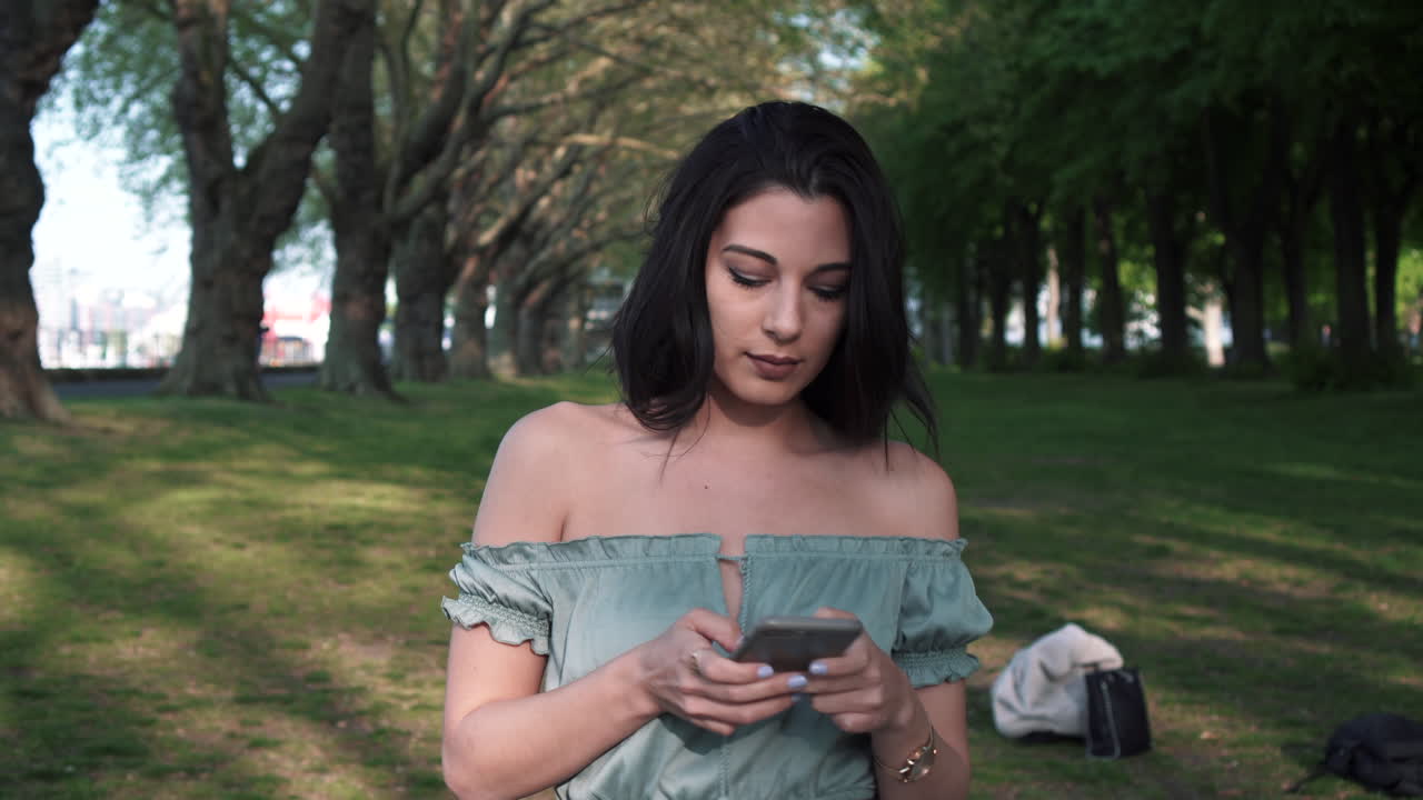 Una giovane ragazza bruna, sicura di sé e attraente, con un top casual, usa il suo smartphone, sorride felice ai messaggi, si guarda intorno mentre cammina in un parco con una fila di alberi sullo sfondo.