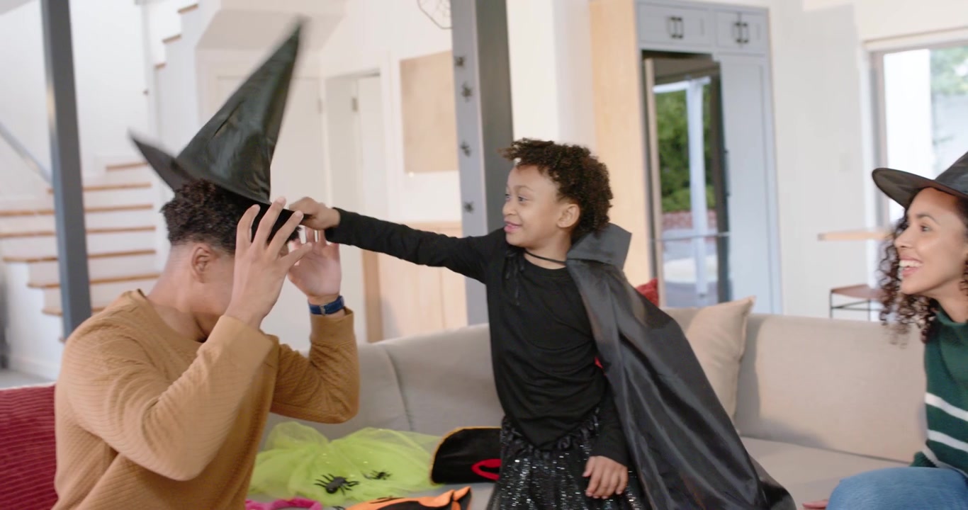 African American family playing witch on couch, woman approaching, fixing cape, child handing hat