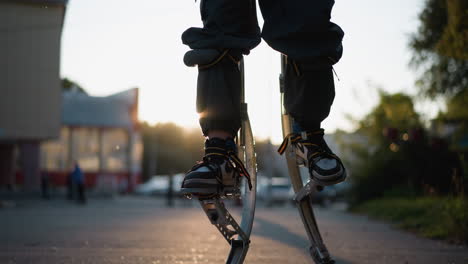 man walking on spring stilts wearing dark pants and sneakers during sunset in suburban outdoor setting. background includes blurred greenery, buildings, and soft sunlight. low-angle view on motion