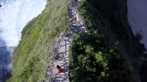 mujer vestida de naranja en la playa de kelingking en la isla de nusa penida indonesia bajando escaleras, toma de vista de rotación aérea