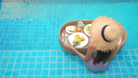 Mujer-Joven-Se-Sienta-En-El-Borde-De-La-Piscina-Con-Bandeja-Flotante-De-Comida