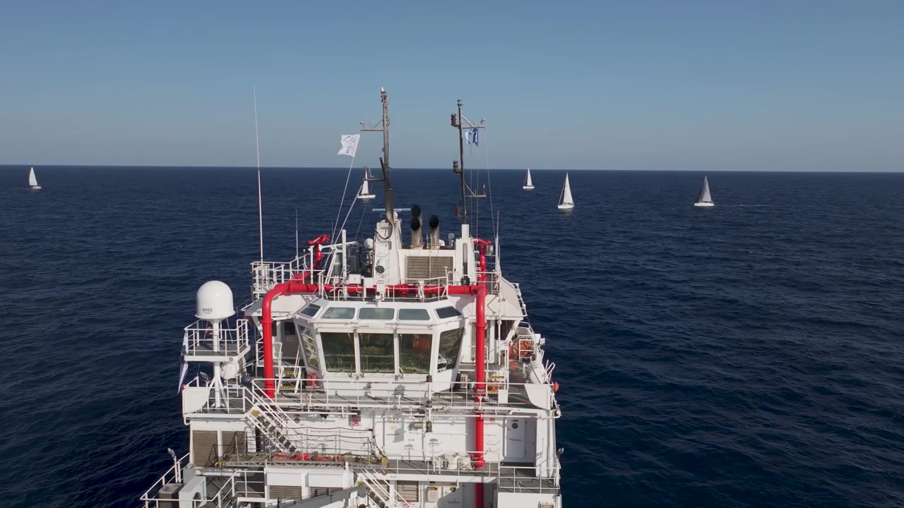 Uma perspectiva elevada captura um barco de serviço branco navegando em meio a iates à vela durante uma regata no mar azul profundo.