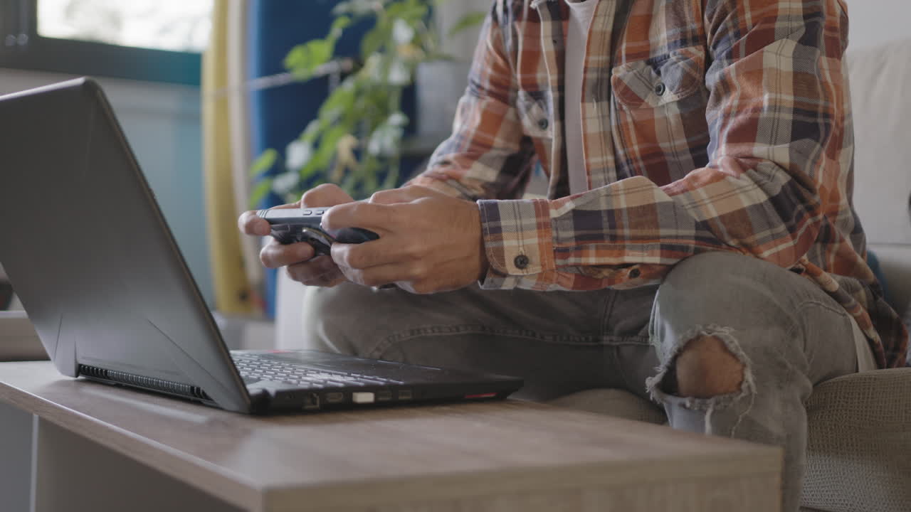 Young adult freelancer working from home in his living room using laptop, is distracting by playing mobile games and watching funny videos content in social networks