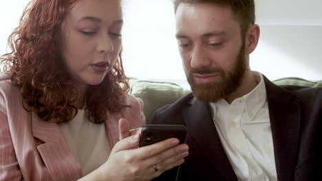 close up of woman and man in formal wear using mobile phone and talking together while sitting on sofa 1