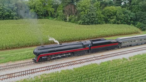 an aerial front to side view of a steam passenger train blowing smoke as it starts to pull out of a small station waiting for passengers to board on a summer day