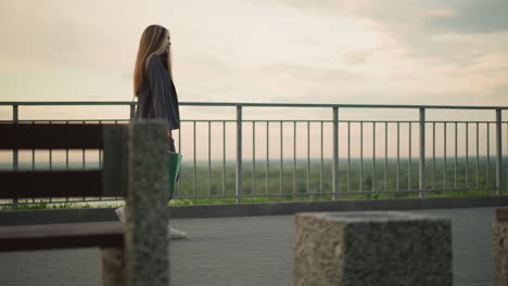 side view of lady in grey clothing walking along iron rail with shopping bag and black handbag, blurred background featuring a stone bench and distant greenery under soft, calm lighting at dawn