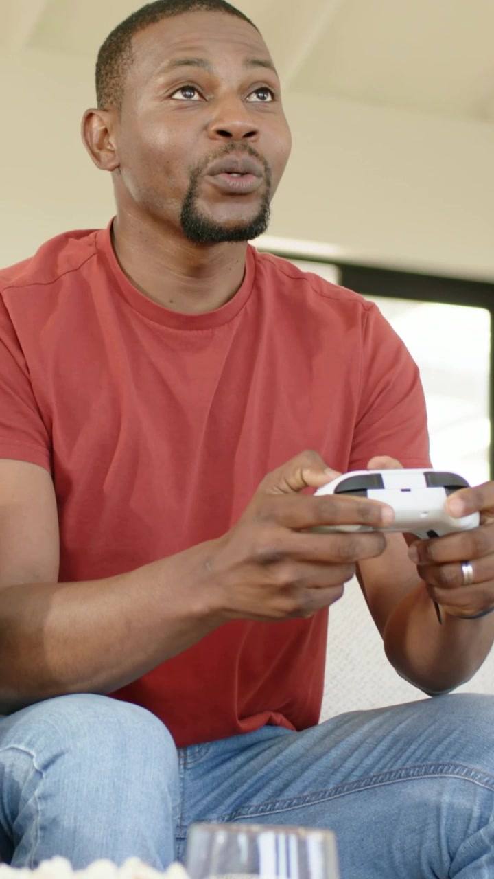 Vertical video: Playing man in red tee reacting to game, leaning on home sofa with pad and popcorn