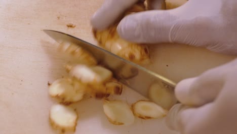 hand with glove using knife to cut galangal into pieces on white chopping board, close up