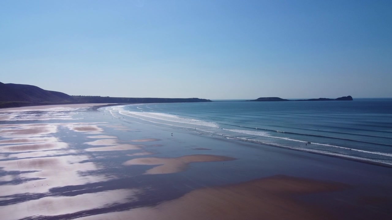 Vista aérea da Praia da Baía de Rhossili com falésia de charneca e o promontório de Worm's Head Causeway silhuetado com o reflexo do sol na areia e na água azul no verão 4K.