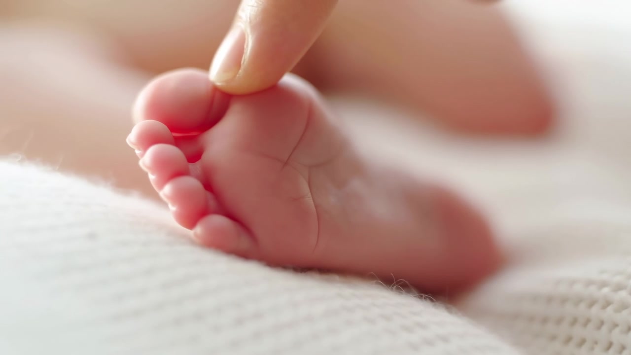 Nærbillede af bevægelsesscene viser voksens fingerspids forsigtigt rører nyfødts tå på et tekstureret strikket tæppe, med let fordybning, bløde hudfolder, varmt naturligt lys og rolig intim pleje