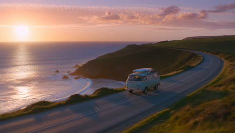 camper van driving on a coastal road at sunset with a surfboard on the roof