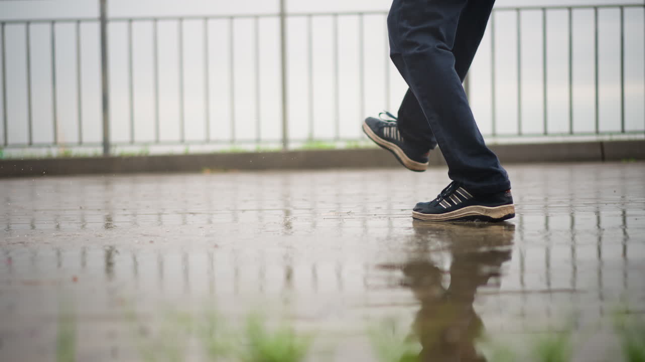 젖은 보도의 다리와 운동화, 발코니 난간을 따라 천천히 조심스럽게 발을 내딛는 통근자, 부드러운 이슬비와 흐린 하늘, 웅덩이에 잔잔한 물결, 배경의 흐릿한 스카이라인과 생각에 잠긴 모습