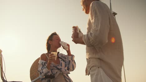 Shot-from-below-against-the-sky,-a-guy-and-a-girl-in-light-beach-clothes-eat-hot-dogs-and-drink-coffee-near-the-sea-in-the-summer.-A-guy-straightens-a-girl's-hair-during-a-date-at-sea