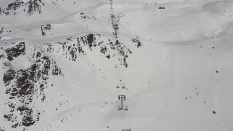 Toma-De-Drones-Inclinada-Hacia-Abajo-De-Un-Telesilla-Muy-Largo,-Una-Enorme-Cadena-Montañosa,-Pistas-De-Esquí-Y-Esquiadores-Montando-Una-Carrera-De-Serpientes-En-Una-Sola-Fila