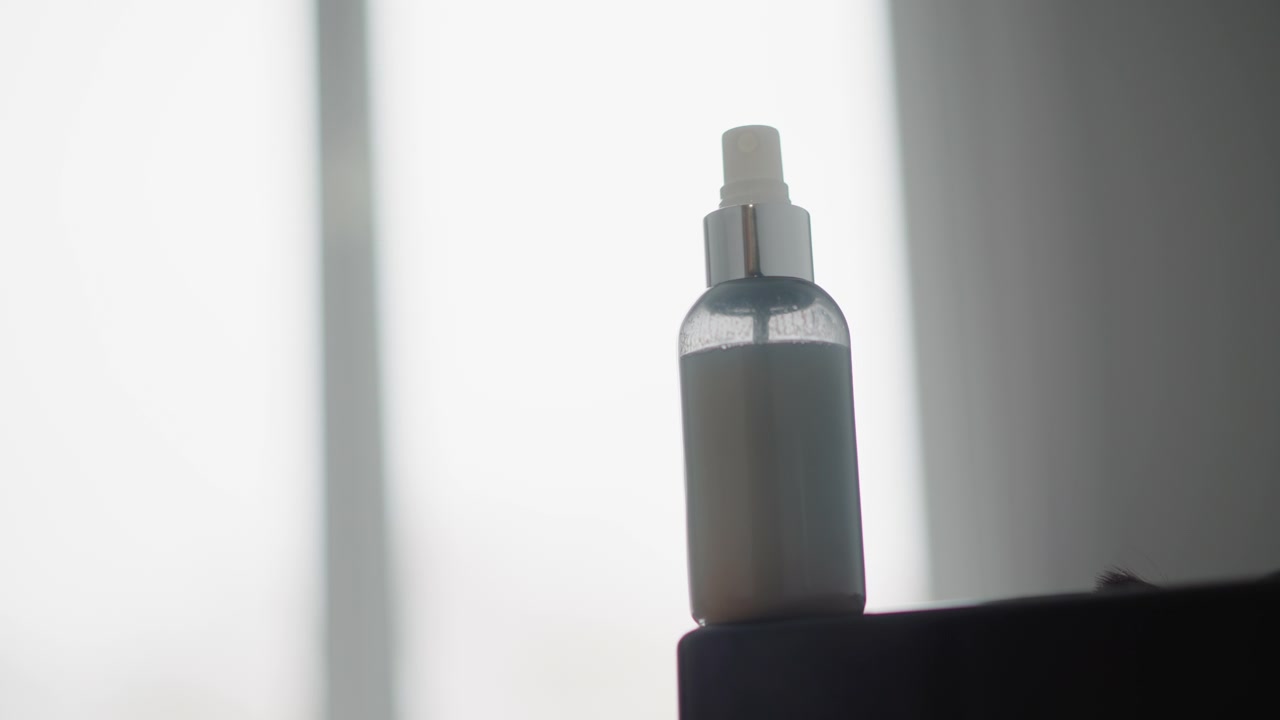 Close up of clear plastic spray bottle with white liquid inside positioned on dark surface. Soft lighting creates gentle shadows around cylindrical object and its metallic cap. Background