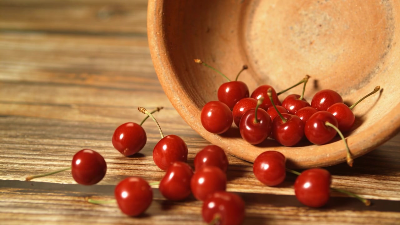Ciliegie rosse che cadono da un vaso di terracotta rovesciato, rotolano e si posano su un piano d'appoggio in legno.