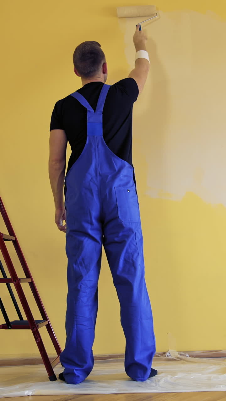 Man in blue overalls painting wall with a roller. Worker decorating room with a new color. Painting services. Rear view. Vertical video