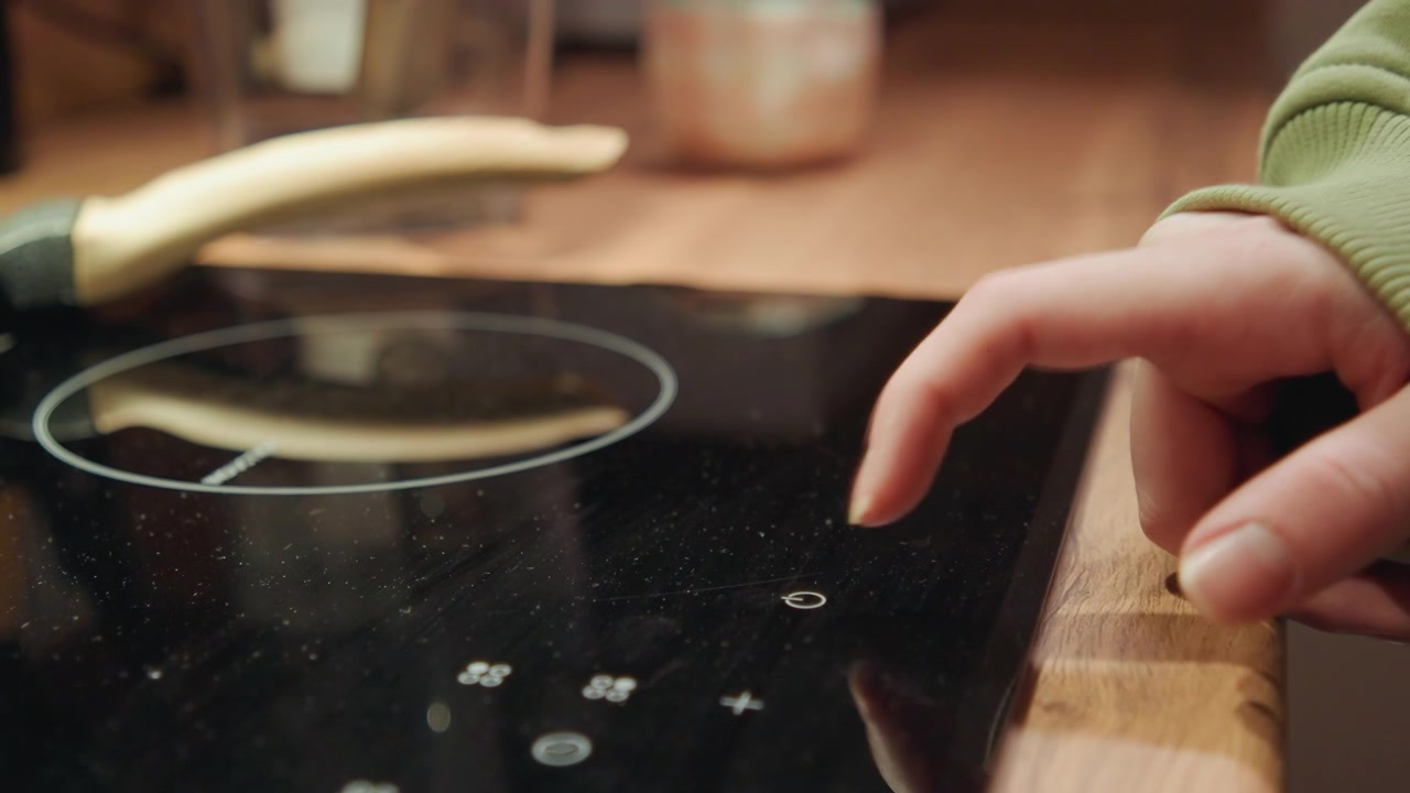 Finger presses illuminated touch controls on glossy black induction cooktop beside metal pot handle on wooden countertop, with warm kitchen lighting, shallow depth, soft reflections, and modern home