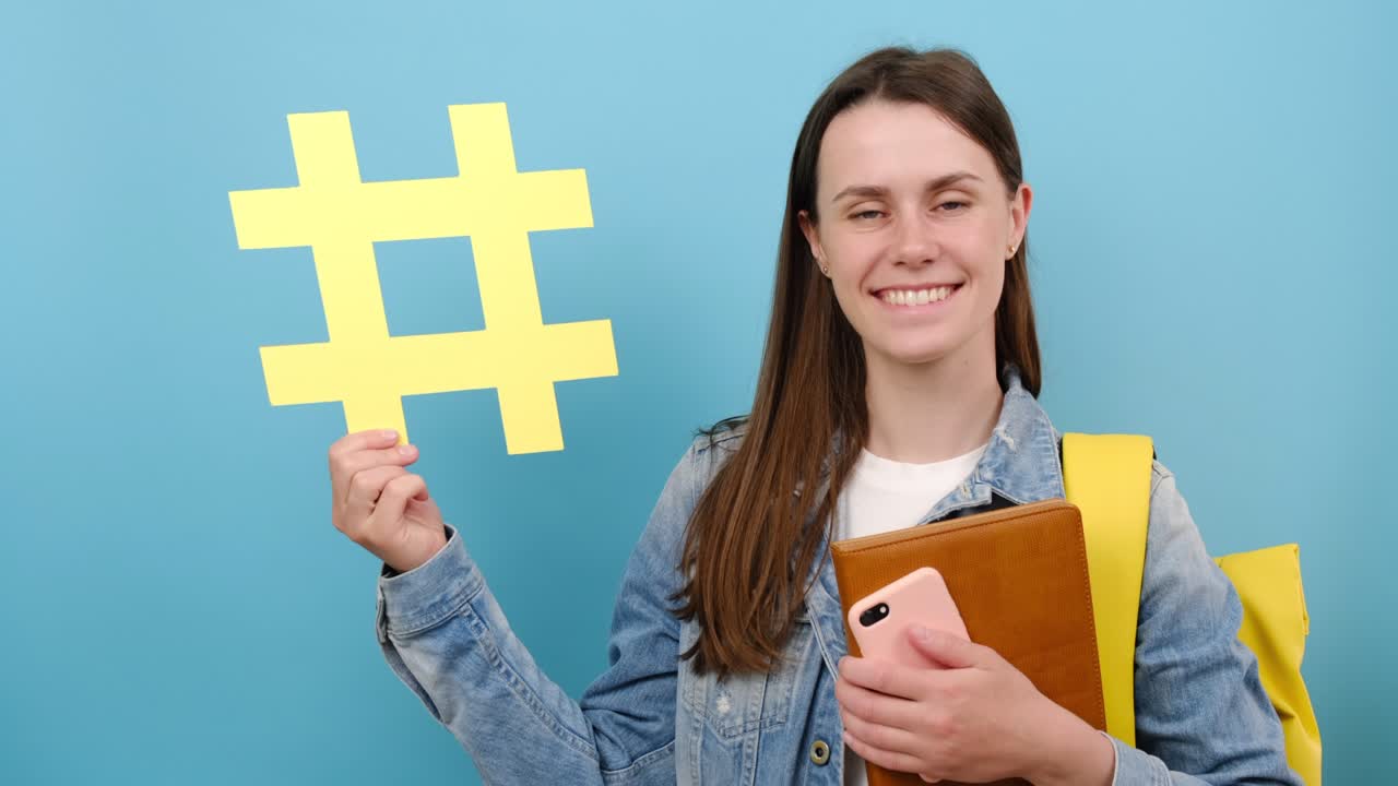 retrato de una atractiva estudiante adolescente sonriente lleva una mochila sostiene libros y muestra hashtag amarillo, modelo sobre pared de fondo de color azul en el estudio con espacio de copia. concepto de monitoreo de redes sociales