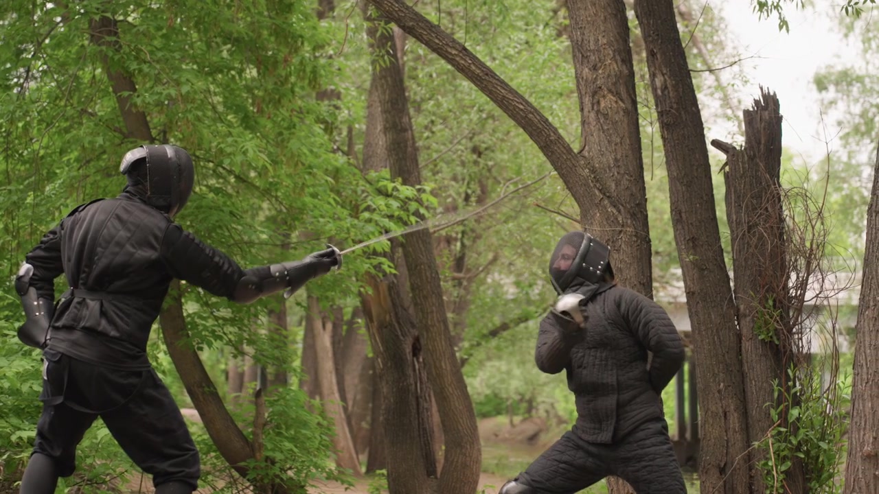 Two people in black protective armor engage in sword fight within dense forest setting demonstrating dynamic movements and techniques. scene features close up views of their stances weapon
