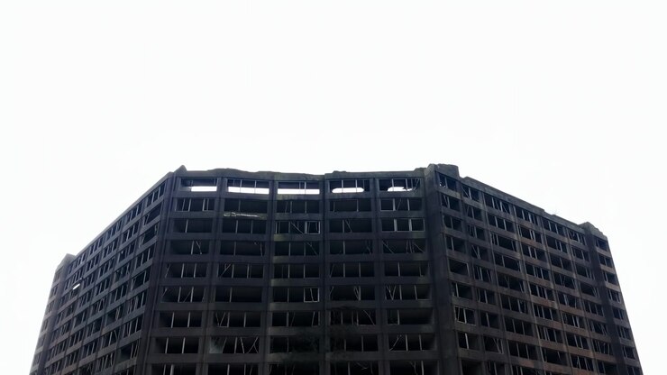 Exterior de edifício escuro de vários andares move-se gradualmente para fora do enquadramento enquanto andares superiores e padrões de janelas permanecem visíveis, mostrando luz do dia suave, linhas simétricas e texturas de superfície urbanas.