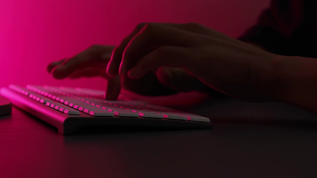 Close side view shows hands actively typing on slim keyboard in dark interior, with vivid magenta lighting, soft blur, glossy reflections, and minimal background creating focused modern tech