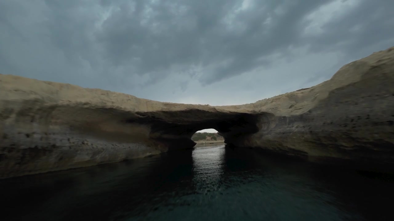 Skimming inches above dark water through the S'Archittu rock arch in Sardinia. FPV drone footage in 4K with brooding storm clouds, pale limestone walls and a glimpse of sunlight beyond.