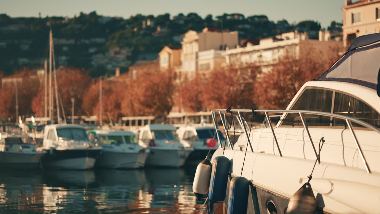 Barche ormeggiate in un porticciolo con edifici residenziali collinari e alberi autunnali sullo sfondo